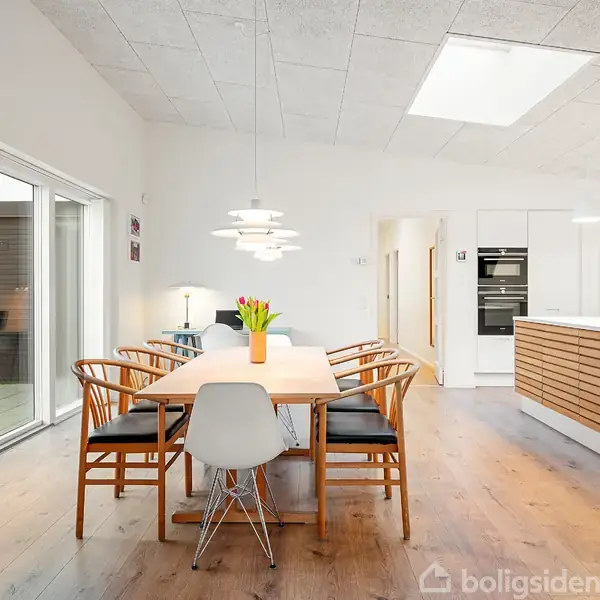 Et spisebord omgivet af stole står i et moderne, lyst køkken-alrum med trægulve. Der er en vase med blomster på bordet, og store vinduer lader naturligt lys ind.