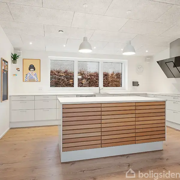 Køkken med træø, skabslåger og moderne apparater, herunder ovn og køleskab. Store vinduer står overfor øen, med to pendellamper. Billede på væggen. Lyst trægulv og hvid loft.