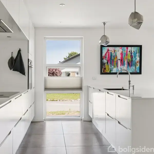 Køkken med hvide skabe og en moderne, farverig kunstplakat på væggen. To pendellamper hænger fra loftet. Store vinduer og en dør giver udsigt til en have.