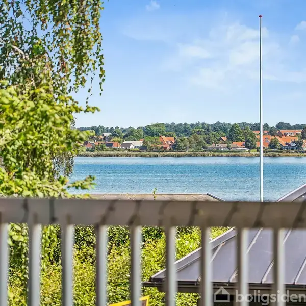 Balkon med hvidt rækværk. Udsigt over en sø med grønne træer og tagene af huse i baggrunden. Klar blå himmel.