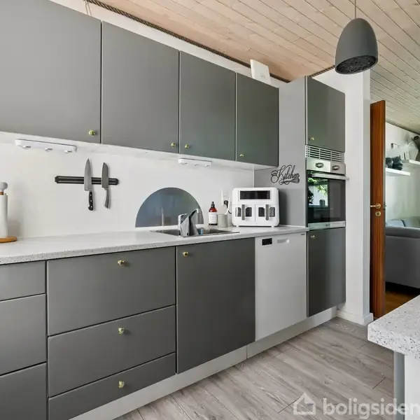Køkken med grå skabe og hvid bordplade. En knivblok hænger på væggen, og der er en kaffemaskine ved siden af indbygget ovn. I baggrunden ses en stue med sofa.