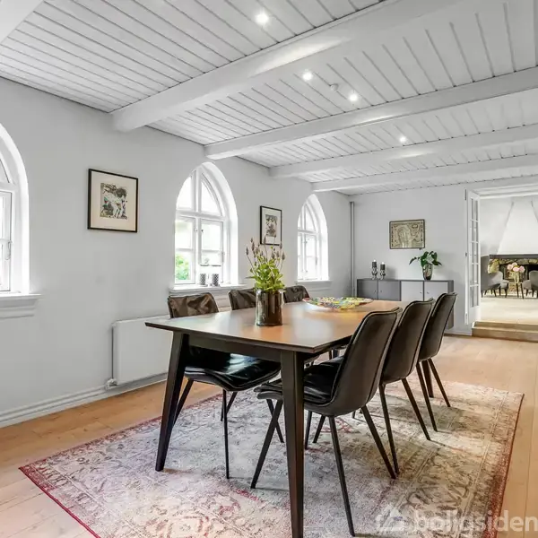 Spisebord med seks stole står i et lyst rum med hvide trævægge og loftsbjælker. Store buede vinduer giver naturligt lys. Et tæppe ligger under bordet, og planter pynter bordet.