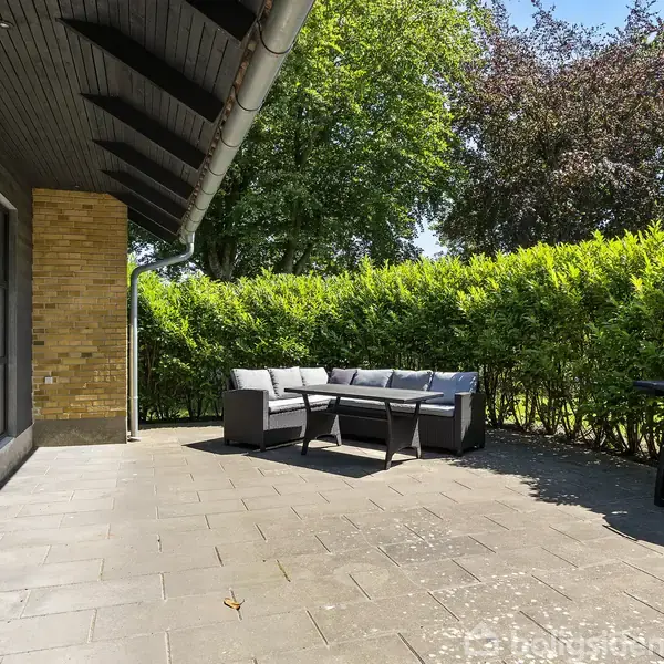 Terrasse omgivet af grønne buske og store træer. Grå loungesofa står til venstre, mens en grill er placeret til højre. Store glasvinduer viser indersiden af huset mod venstre.