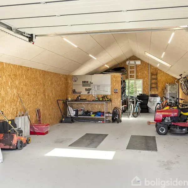 En garage med åbne døre indeholder en rød havetraktor, cykler ophængt på væggen, værktøjer på hylder og en arbejdsbænk. Brædder dækker væggene, og lysstofrør hænger fra loftet.