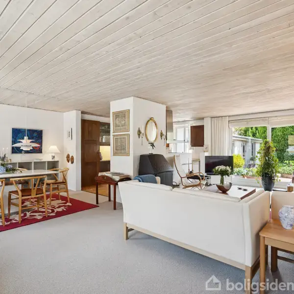 Stue med lyse trægulve og hvidt loft, der har røde dekorationer. Indrettet med spisebord til otte personer, hvide sofaer og udsigt gennem store vinduer til en have.