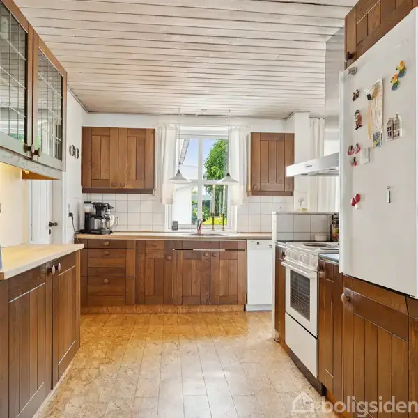 Køkken med brune træskabe, hvidt køleskab med magneter, komfur og kakkelbordplade. Lyse trælofter og gulv. Vindue med udsigt til grønne træer. Kaffeapparat på bordet.