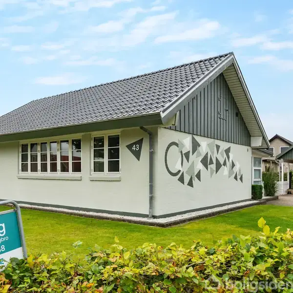 Hus med sort tegltag og hvide vægge, nummer 43, står i en have. Skilt foran huset har teksten: "Til salg, danbolig Bjerringbro, 86 68 12 48."