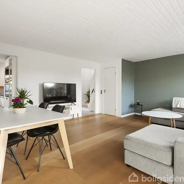 En stue med en grå hjørnesofa og et hvidt sofabord mod en grøn væg. Et spisebord med sorte stole står ved siden af, og et TV hænger på væggen. Der er adgang til et køkken.