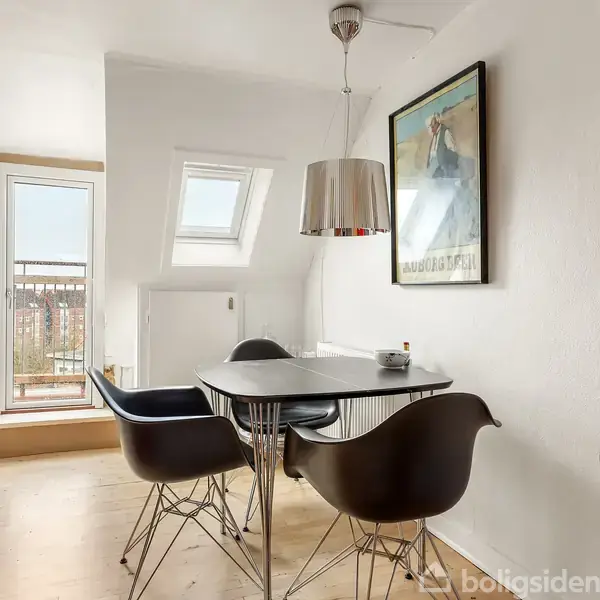 Et rundt spisebord med fire sorte stole står i et lyst rum. En stor lampe hænger over bordet. Rummet har et skråvindue og en dør til en balkon. En plakat hænger på væggen.