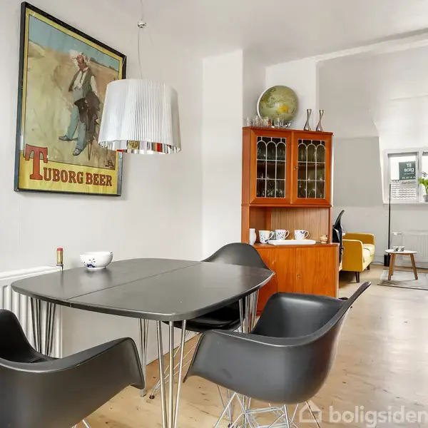 Et sort bord med sorte stole står i en lys stue med et Tuborg-ølplakat på væggen, en lampe hænger over bordet, og en træskænk står i baggrunden.