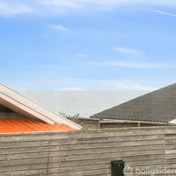 To tage med skrånende røde og grå overflader er synlige. I baggrunden ses havet under en blå himmel med skyer.