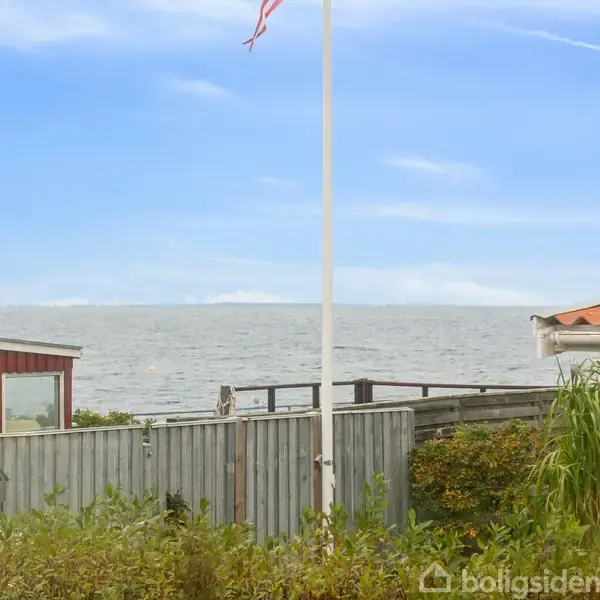 Flagstang står stille, omgivet af buske og hegn, mod en baggrund af blåt hav og himmel; røde og gule husgavle ses til siderne.