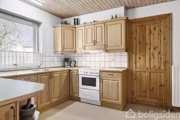 Køkken med træskabe og fliser på væggen, komfur centralt placeret. Vindue med udsigt til have. Trædør til højre for skabene. På skabet står dekorative figurer.