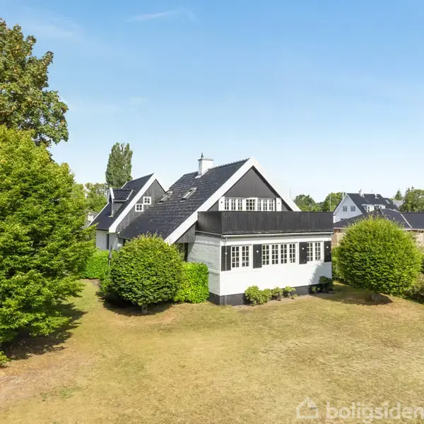 Et hvidt hus med sort tag står på en stor plæne omgivet af buske og træer i et villakvarter. Himmel er blå med klart vejr.