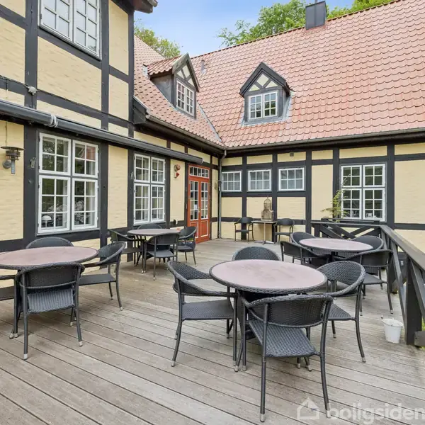 Havemøbler står på en terrasse, der er del af et bindingsværkshus med gule vægge og rødt tegltag. I baggrunden er der en frodig have med træer og buske.