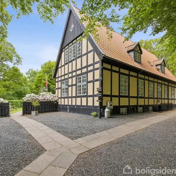 Hus med bindingsværk står stille omgivet af grønne træer og buske. Grå grusbelægning fører til indgang. Husets tag er rødt, og der ses blå himmel.