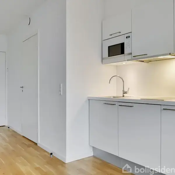 Køkkenområde med lyse, moderne skabe og indbygget mikrobølgeovn placeret over en vask. Håndvask på bordplade. Sammenhængende trægulv udstrakt mod to hvide døre i baggrunden.