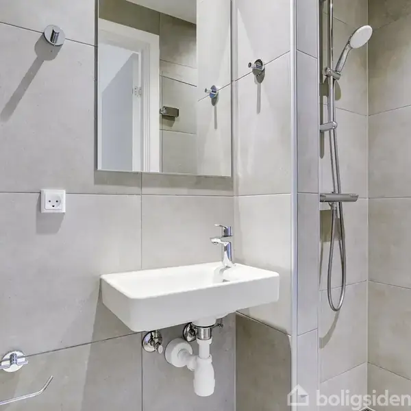 Håndvask monteret på væggen, omgivet af store, grå fliser. Over vasken hænger et spejl. Bruseren er til højre med metalarmatur. Væggen har en stikkontakt og toiletrulleholder.