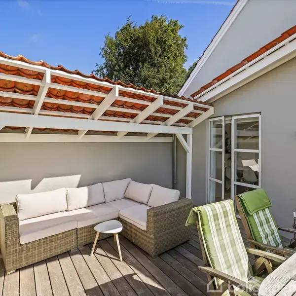 En udendørs terrasse med sofa under en halvtag. To afklappede stole med stribede håndklæder ved et bord. Blåt himmel og træ i baggrunden.