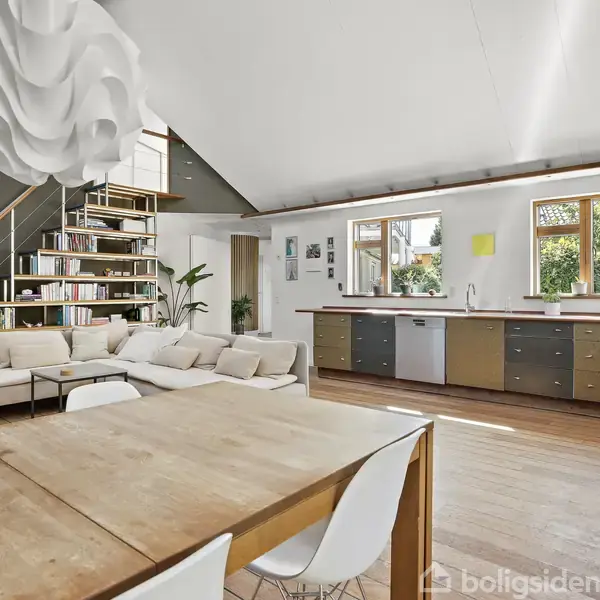 Lys stue med træmøbler og sofa. Reol langs væggen ved trappe. Stor, hvid loftlampe hænger ned. Køkken med vinduer og grøn udsigt i baggrunden. Ingen tekst tilgængelig.