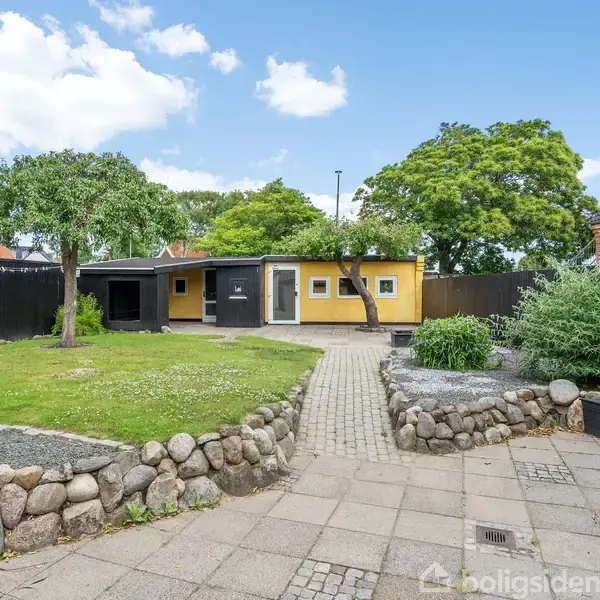 En gul bygning med fladt tag står i baghaven omgivet af grønne træer og græs. En brolagt sti fører fra terrassen til huset, med en stenmur på siderne.