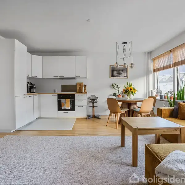 Stue med sofa og sofabord, set mod åbent køkken med hvide skabe; spisebord med blomster ved vindue med træpersienner, skaber en lys og moderne atmosfære.