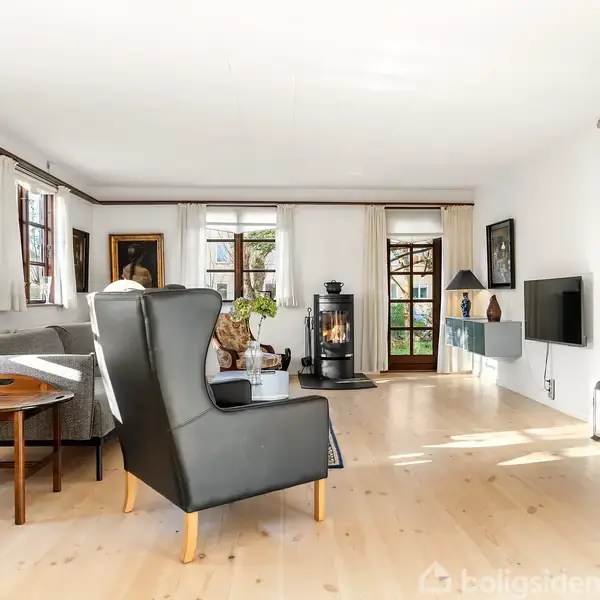 En moderne stue med sorte lænestole foran et brændende ildsted. Store vinduer med hvide gardiner slipper lys ind. På væggen hænger en fladskærm. En åben dør vises til højre.