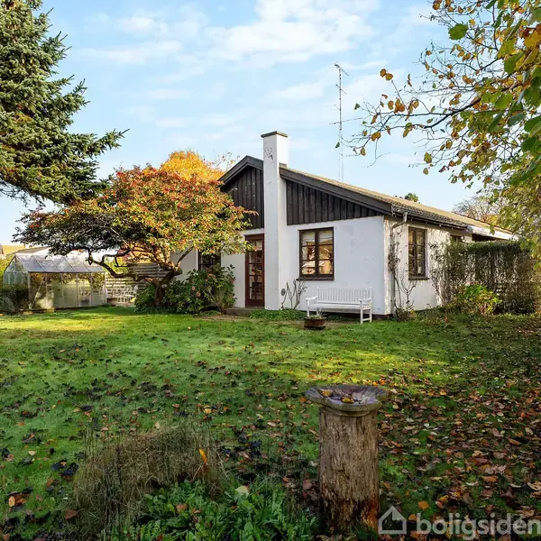 Hus med hvide vægge og mørkt tag står i en grøn have. Træer med efterårsløv omgiver huset, og en hvid bænk er placeret foran.