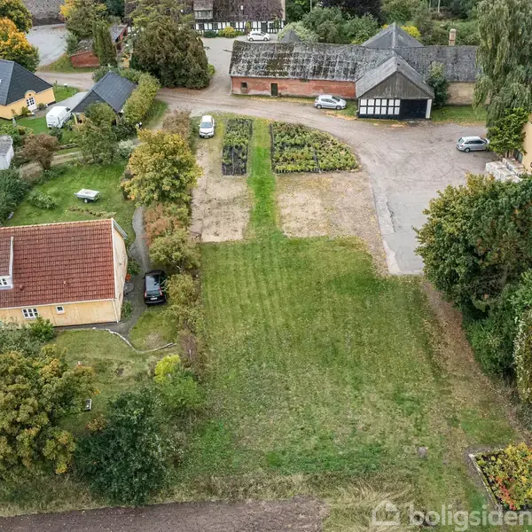 Græsareal med grusvej fører til flere bygninger, herunder et gult hus med rødt tag. Omgivet af træer, haver og landejendomme, ses også parkerede biler på grusplads.