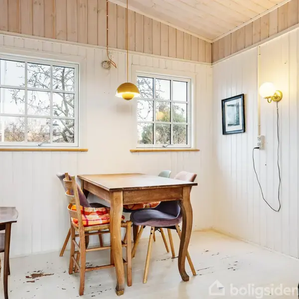 Et træbord med stole står i et lyst rum med træpaneler. To vinduer lader sollys komme ind. Der hænger en lampe over bordet, og en væglampe lyser op til siden.