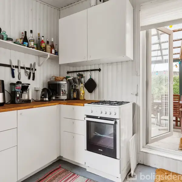 Køkken med hvide skabe, en ovn, køleskab og køkkenudstyr på bordpladen. Gad døre fører ud til en terrasse med havemøbler. Væggene er hvidmalet paneler. Gulvtæppe ligger på gulvet.