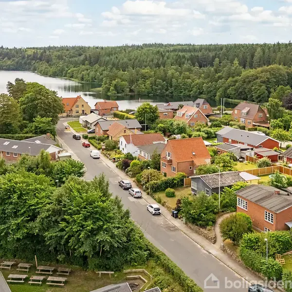 Et villakvarter omgivet af frodig skov med en sø i baggrunden. Forskellige huse ligger langs en vej, og himlen er let overskyet.