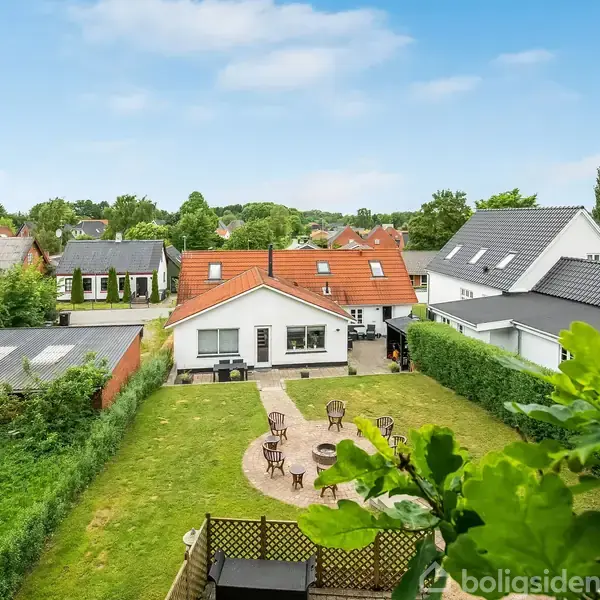 Et hvidt hus med rødt tag står i en have med terrasse og stole. Omgivet af andre huse og grønne træer under en blå himmel.