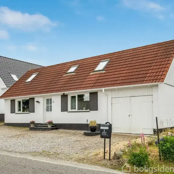 Et hvidt hus med rødt tag står ved en vej. Foran er der en grusindkørsel og en lille sort postkasse. Til højre ses grønne buske og træer.