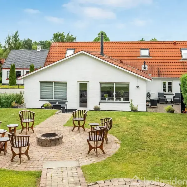 En hvid villa med rødt tag står i en grøn have. En rund brolagt terrasse med stole og bord omkranser en ildsted i midten.