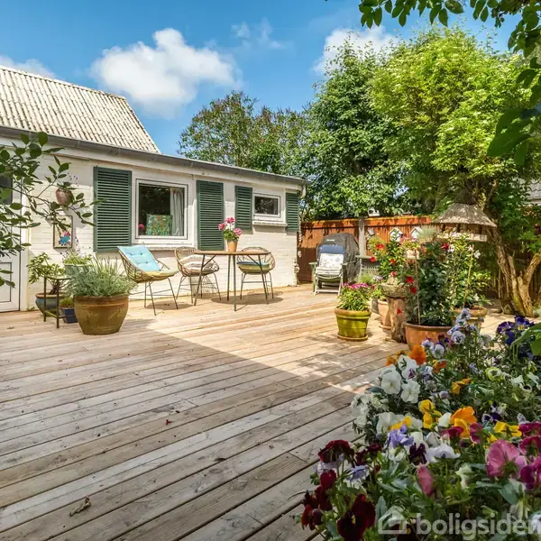 Et træterrasseområde med blomsterkrukker, cafébord og stole foran et hvidt hus med grønne skodder. Terrassen er omgivet af frodig beplantning og et træhegn.