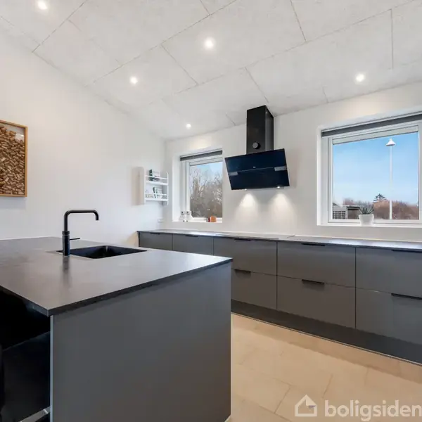 Køkken med mørkegråt køkkenbord, sort emhætte over kogepladen, lyse vægge og loft med indbyggede spots. To store vinduer giver udsigt til træer udenfor. Mørke stole ved bord.