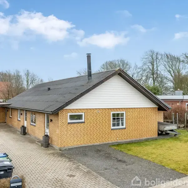 Et gult murstenshus står stille på en grusbelagt indkørsel med grøn have ved siden af. Huset er omgivet af træer og nabobygninger under en blå himmel med få skyer.