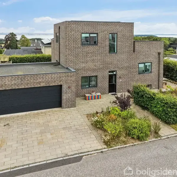 Moderne to-etagers murstenshus, står stille på en flisebelagt indkørsel. Huset er omgivet af velholdt have og hæk. Blå himmel og nabolagets huse ses i baggrunden.