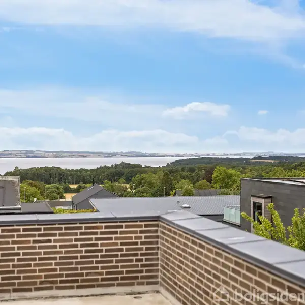 En murstensmur tilhørende en tagterrasse med udsigt over et skovområde og en fjord i baggrunden, under en blå skyet himmel.
