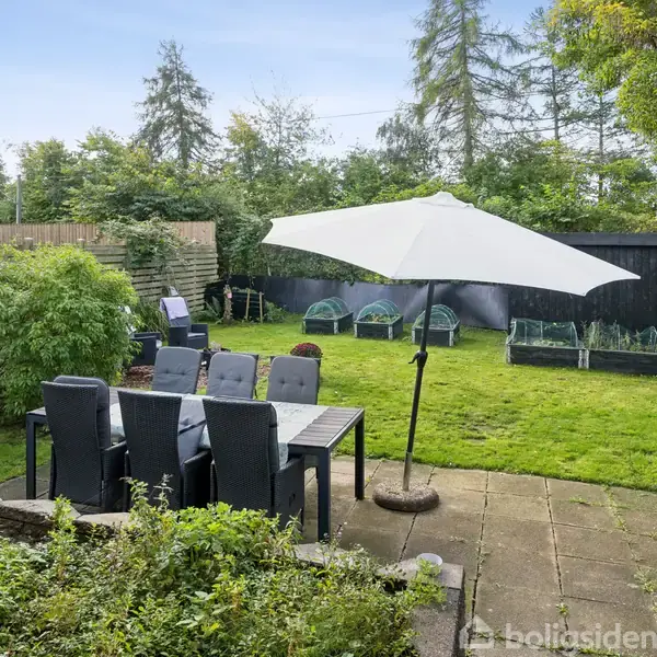 Et hus med stor have, hvor et spisebord med parasol står på en terrasse. Græsplænen er omgivet af hegn og buske, med flere siddepladser og træer i baggrunden.
