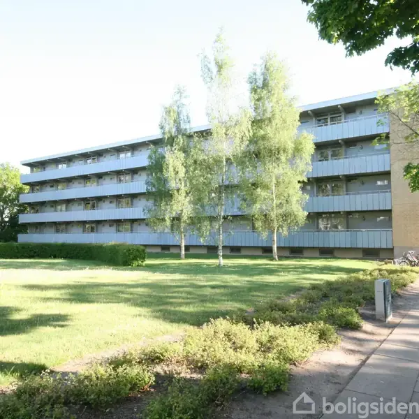 En fleretagers boligbygning med balkoner omkranset af grønne træer. Foran bygningen er der en velplejet græsplæne og buskads. Solrig dag med skygger på fortovet.