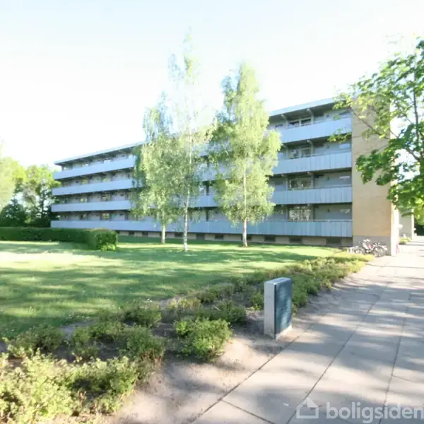 Et større lejlighedskompleks med flere etager står omkranset af grønne træer og en velholdt græsplæne. Fortov og parkerede biler ses til højre i billedet.