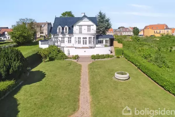 Stort, hvidt hus med blå tagsten står på en velplejet græsplæne. Huset har flere vinduer og en terrasse. Haven er omkranset af grønne buske og en murstenssti leder op til huset.