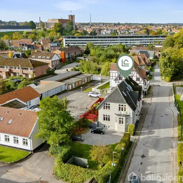 En grå, stor bygning står centralt med parkeringsplads foran. Den er omgivet af mindre huse og grønne træer i et boligområde. I baggrunden ses en kirke og flere træer.