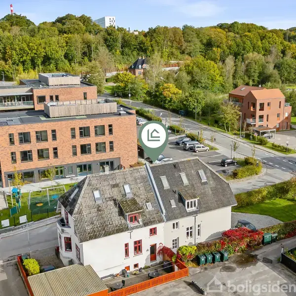 En hvid bygning med skråtag står centralt, omgivet af røde murstensbygninger og grønne områder. I baggrunden er der skovbevoksede bakker. En vej løber foran bygningerne.