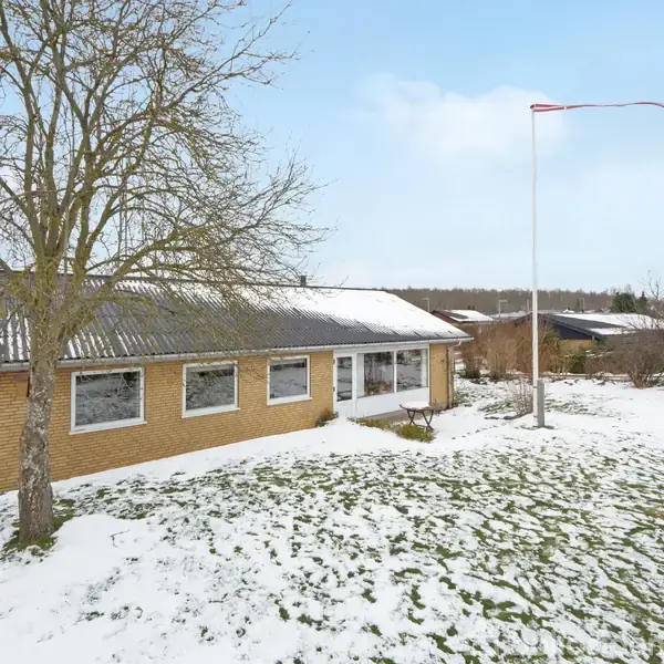 Et enfamiliehus i gule mursten står stille omgivet af en snedækket have. Et træ uden blade og to flagstænger ses på grunden under en blå himmel.