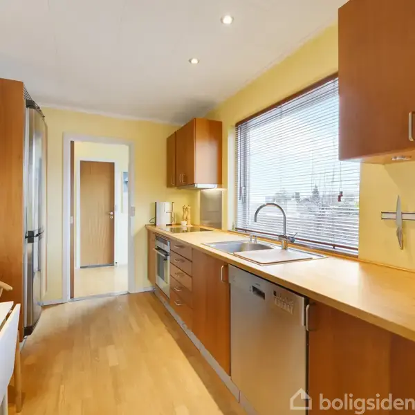 Køkken med træskabe og opvaskemaskine ved et stort vindue. Skarpe knive er monteret på væggen over træbordpladen. Bord og stole står til venstre, dør forrest mod gangen.