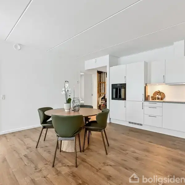 Et moderne køkkenalrum med hvidt køkkenskab og en rund spisebord med grønne stole. Der er lyse trægulve og en åben dør til et soveværelse med en seng.