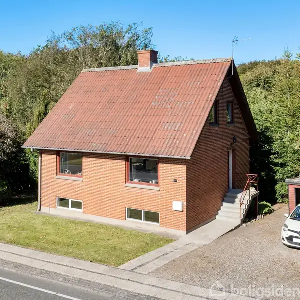 Et rødt murstenshus med skråt tag står ved siden af en lille parkeringsplads med en hvid bil, omgivet af træer og buske.